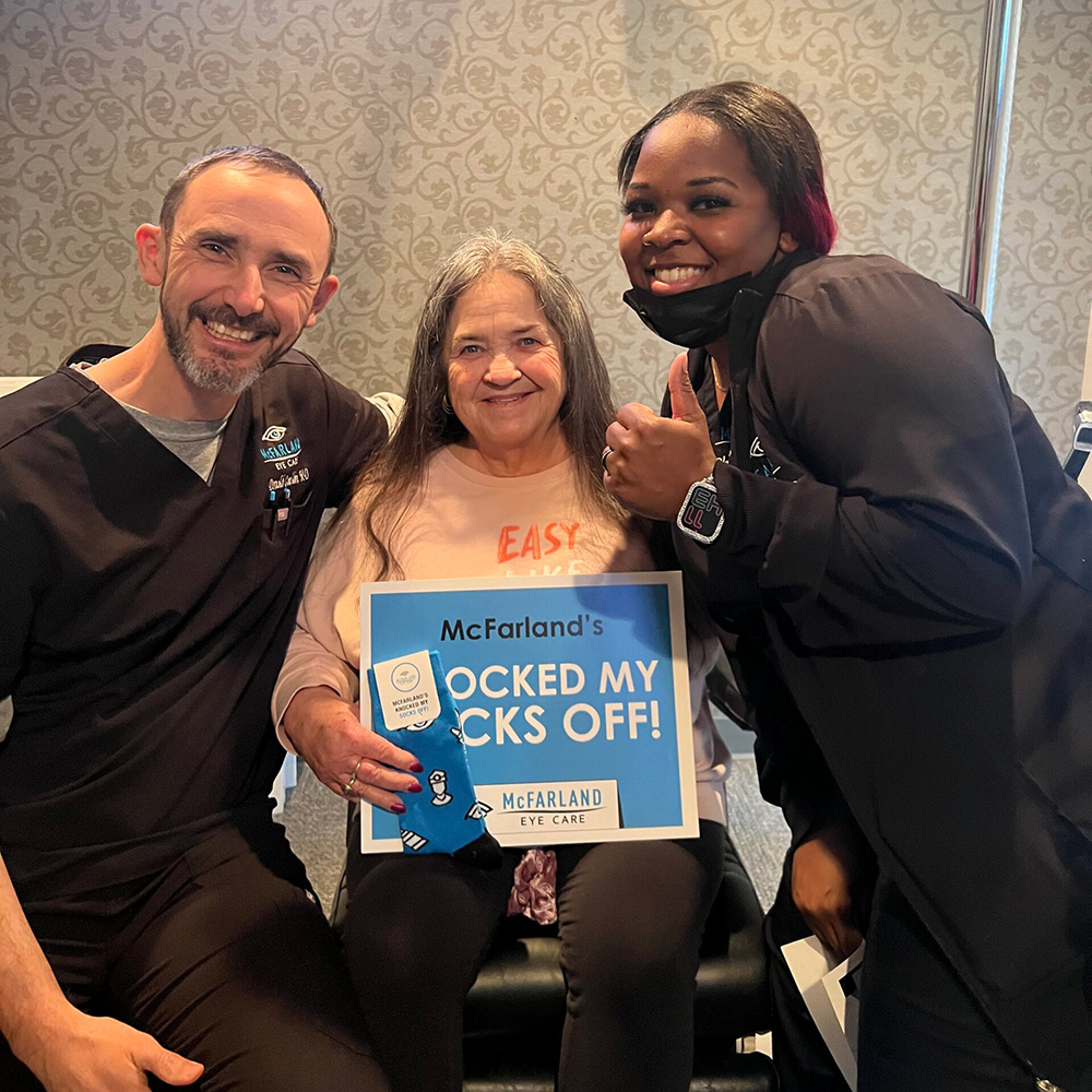 Happy cataract surgery patient posing with two McFarland Eye Care team members while holding a “Knocked My Socks Off!” sign.